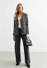 Zwart-wit gebreide blazer met franjes, gedragen over een witte top. Gecombineerd met hoge taille, brede donkere jeans en een kleine zwarte handtas.