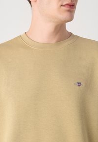 Beige katoenen sweatshirt met een ronde hals, geribde manchetten en een klein geborduurd logo op de linkerkant van de borst. Soepele textuur, casual fit.