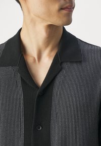 Homme portant un polo en maille noire avec des rayures verticales texturées grises et noires, boutonné au niveau de la poitrine, regardant vers la droite.