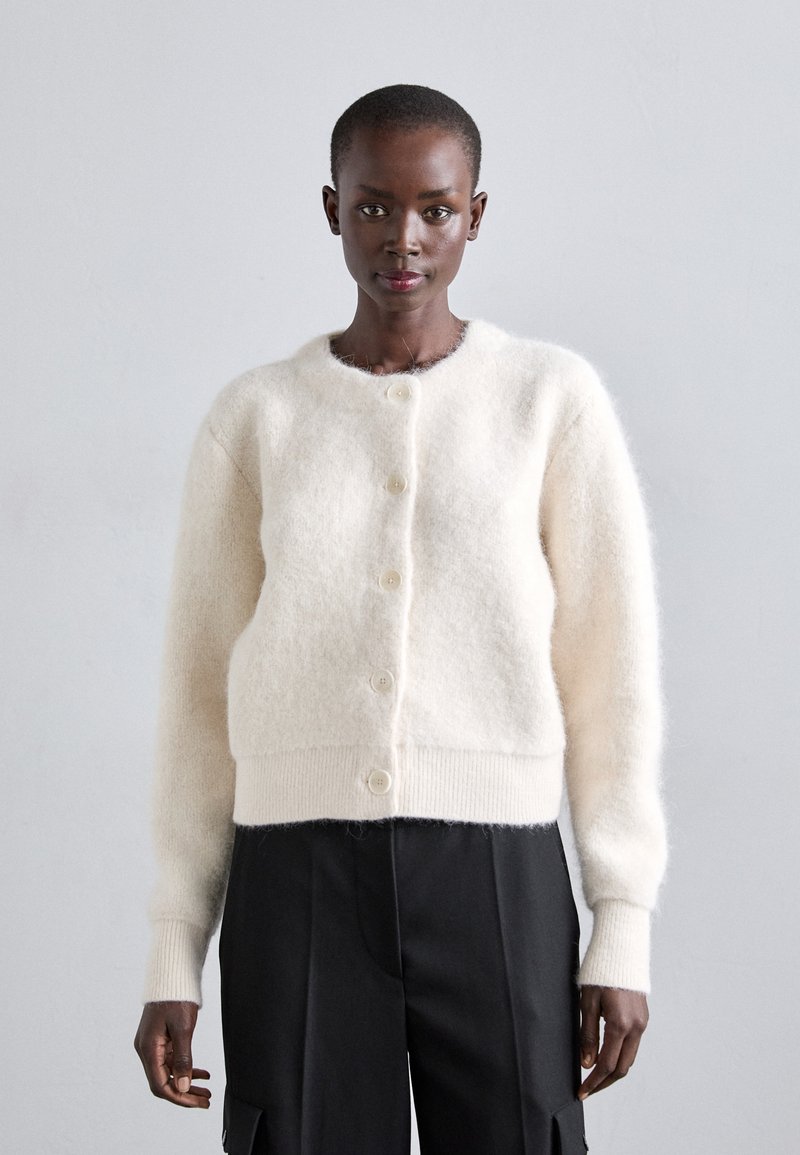 Una persona di colore che indossa un cardigan bianco peloso con bottoni e pantaloni larghi neri, in piedi davanti a uno sfondo grigio chiaro uniforme.