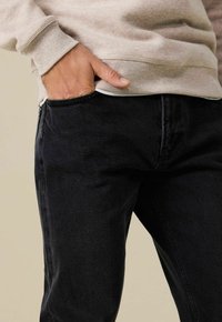 Jean en denim noir à coupe slim, avec une poche avant, des coutures visibles et une texture douce. Porté avec un sweat-shirt beige clair.