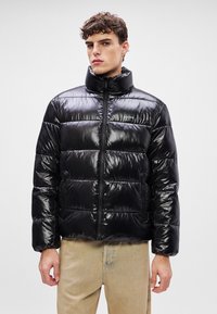 Μαύρο μπουφάν puffer με γυαλιστερή, καπιτονέ επένδυση, ψηλό γιακά και μπροστινό φερμουάρ. Διαθέτει πλαϊνές τσέπες και διακριτικό λογότυπο στο στήθος.