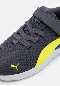 Sneaker grigio scuro con tomaia in mesh, dettagli gialli e suola flessibile. Presenta lacci piatti e una fascia per una vestibilità sicura. Logo PUMA visibile.