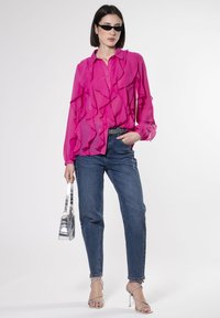 Blusa fucsia con ruffles, maniche lunghe e chiusura con bottoni. Abbinata a jeans blu a vita alta, tacchi a stiletto argentati e una borsa metallizzata.