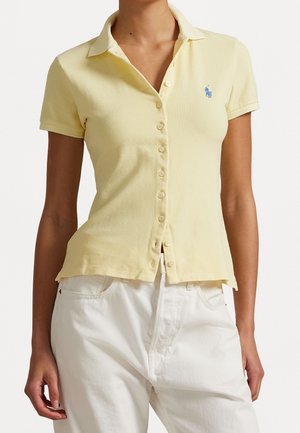 Femme portant un polo jaune pâle à manches courtes boutonné avec un logo brodé bleu et un pantalon blanc.