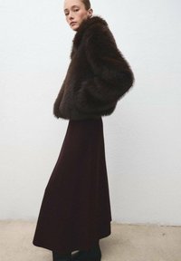 Chaqueta de pelaje sintético marrón con un cuello alto, combinada con una falda larga burdeos. La chaqueta tiene bolsillos laterales y una apariencia texturizada.