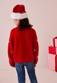 Maglione rosso soffice con maniche lunghe e motivi a stelle dorate. Abbinato a un cappello di Babbo Natale rosso con bordo in pelliccia bianca. Jeans blu.
