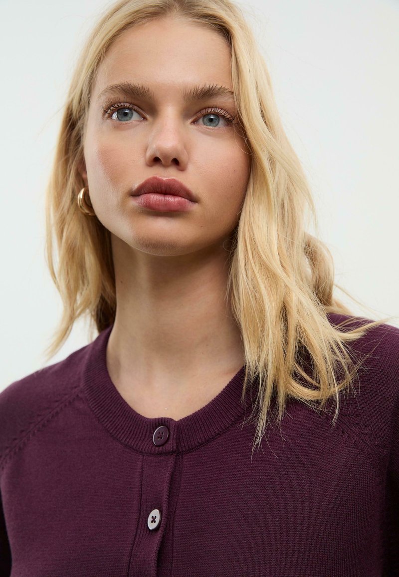 Cardigan a maglia bordeaux con scollo rotondo, due bottoni e maniche raglan. Tessuto liscio con dettagli di cucitura sottili.