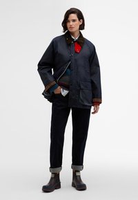 Ναυτικό αδιάβροχο μπουφάν με καφέ κοτλέ γιακά, δύο μπροστινές τσέπες, κλείσιμο με κουμπί, φορεμένο πάνω από πολύχρωμες πλεκτές μπλούζες.