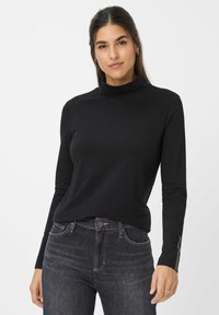 Frau mit dunklen Haaren, die einen schwarzen Langarmrollkragenpullover und dunkle Jeans trägt, vor einem schlichten hellen Hintergrund steht.