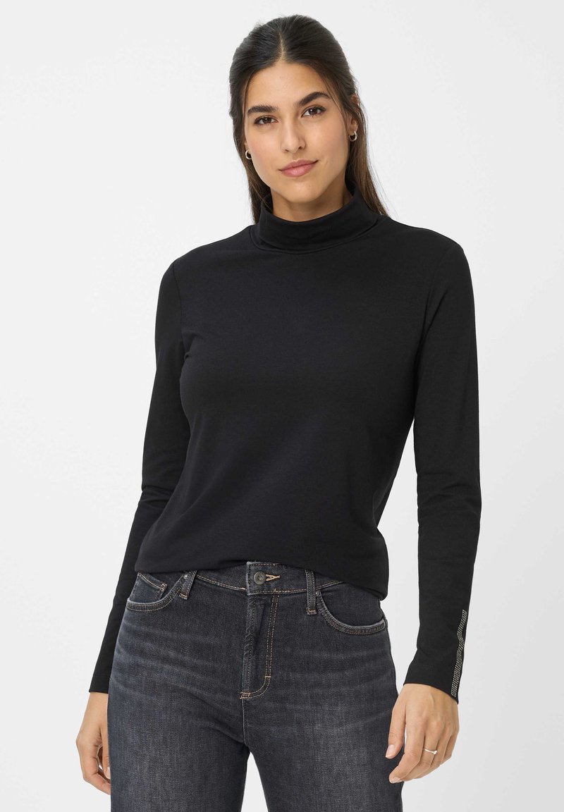 Frau mit dunklen Haaren, die einen schwarzen Langarmrollkragenpullover und dunkle Jeans trägt, vor einem schlichten hellen Hintergrund steht.