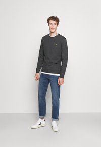 Felpa grigia in misto cotone, con scollo rotondo e logo giallo sul petto. Abbinata a jeans blu e sneakers bianche con dettagli neri.