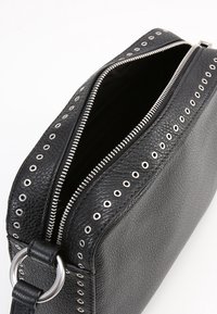 Svart läder crossbody-väska med texturerad yta, silverfärgade nitar längs dragkedjan och en metallring som accent på axelremmen.