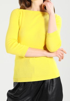 Personne portant un pull jaune vif à manches longues avec des manches trois-quarts et un pantalon noir ample, posant les bras croisés.