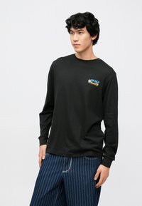 Zwart T-shirt met lange mouwen van katoen, voorzien van kleurrijke tekstborduursels. Draagt losse, gestreepte spijkerbroeken.