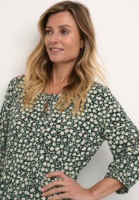 Žalia gėlių suknelė su apvaliu iškirpimu ir rakto skylutės detale, puošta baltais ir rožiniais ramunėlėmis, sukrautu liemeniu ir trijų ketvirčių ilgio rankovėmis.