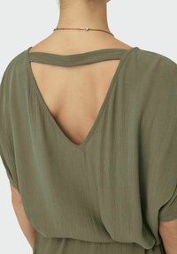 Olivengrünes Kleid aus einem weichen, strukturierten Stoff. Verfügt über einen tiefen V-Ausschnitt hinten und breite Ärmel, ergänzt durch eine zarte goldene Kettenhalskette.