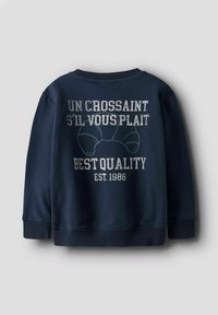 Sweatshirt bleu marine avec des poignets et un ourlet côtelés ; présente un design de texte à l'avant avec des graphiques de croissants, indiquant "Un croissant s'il vous plaît" et "Meilleure qualité 1986."