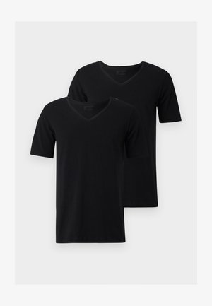 Mand stående barfodet iført sort kortærmet V-hals skjorte og lysegrå joggingbukser mod en ensfarvet hvid baggrund.