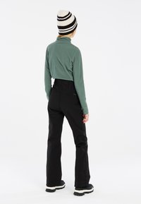 Pull en polaire vert et pantalon de ski noir taille haute, associés à un bonnet rayé noir et blanc, vus de dos.