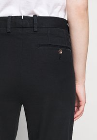 Svarta byxor med en slät yta, med en bakficka med knappdetalj och bälteshällor längs midjan, som visar en skräddarsydd design.