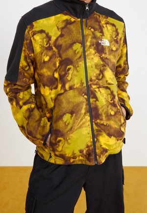 Homme portant une veste polaire North Face jaune et marron à motifs, avec des épaules noires, une fermeture éclair noire et les mains dans les poches latérales.