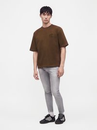 Νέος άνδρας στέκεται φορώντας ένα καφέ oversized μπλουζάκι, ανοιχτό γκρι στενά τζιν, λευκές κάλτσες και μαύρα αθλητικά παπούτσια με λευκές ρίγες.