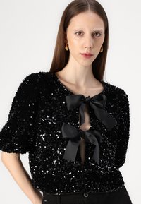 Crna sequin majica s napuhanim rukavima, koja ima crnu traku za vezanje na izrezu i blago otvoreni prednji dio. Glatka tekstura tkanine.