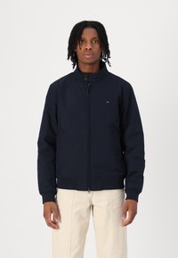 Veste bombardier bleu marine avec un col haut, fermeture éclair sur le devant et poches latérales. Comprend des poignets et un ourlet côtelés, avec un petit logo sur la poitrine.