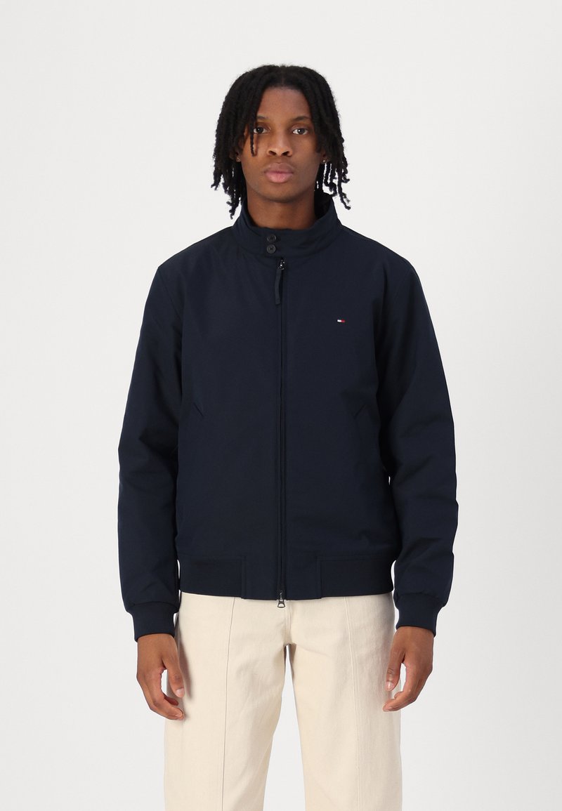 Veste bombardier bleu marine avec un col haut, fermeture éclair sur le devant et poches latérales. Comprend des poignets et un ourlet côtelés, avec un petit logo sur la poitrine.