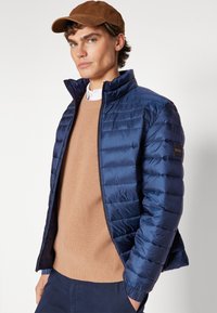 Chaqueta acolchada azul marino con paneles acolchados horizontales, cuello tipo elevado y un parche con logo en el hombro. Llevada sobre un suéter beige.