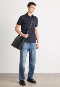 Juodas oda messenger krepšys su reguliuojama juostele, dėvimas su tamsiai mėlynu polo marškinėliu ir šviesiai mėlynomis džinsinėmis kelnėmis. Kasdienis stilius ir tvirta konstrukcija.