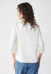 Blouse blanche avec manches bouffantes trois-quarts, tissu lisse et encolure arrondie ; associée à un jean bleu en denim.