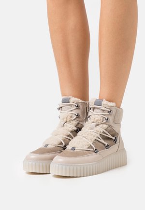 Botas de nieve altas de color beige con detalles de borrego, paneles texturizados, punta redonda y una suela gruesa translúcida estriada. Diseño con cordones que incluye herrajes metálicos.