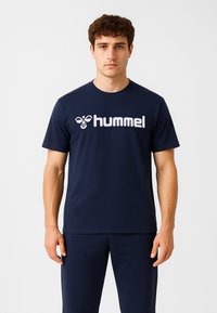 Młody mężczyzna o krótkich, kręconych brązowych włosach, ubrany w granatowy t-shirt i spodnie, stojący na tle gładkiej, białej ściany.