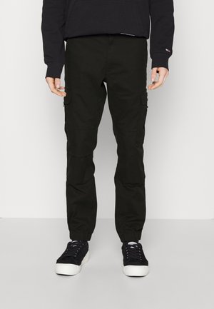 Pantaloni cargo verde scuro con polsini elastici, tasche frontali e tasche cargo laterali. Il materiale sembra resistente e leggermente strutturato.