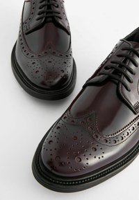 Burgundy színű bőr brogue cipők díszes perforációkkal, hegyes orral és fekete gumitalppal. Klasszikus fűzős kialakítással és sima felülettel rendelkezik.