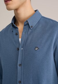 Camisa azul de botones con un patrón texturizado, con un pequeño logo bordado de un pájaro en el pecho y botones oscuros en la tapeta.