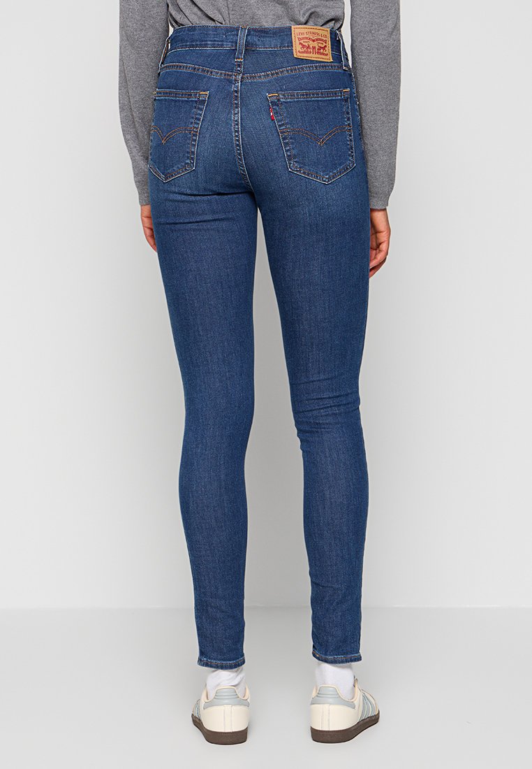 Schlanke blaue Jeans mit mittlerer Leibhöhe, ausgestattet mit einem Lederetikett, zwei Gesäßtaschen und einer glatten Textur. Sichtbare lässige Sneaker.