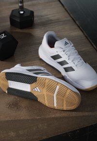 Chaussures de sport blanches Adidas avec des bandes noires reposant sur un sol en bois près de deux haltères noirs.