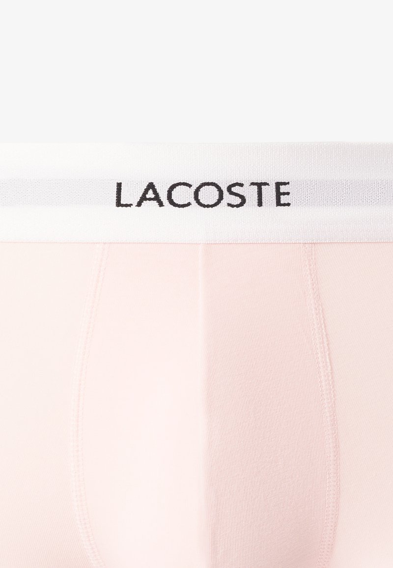 Lacoste Herren-Unterhosen aus zartrosa Stoff mit einem weißen elastischen Bund, der schwarzen Logotext und subtile Nähdetails aufweist.