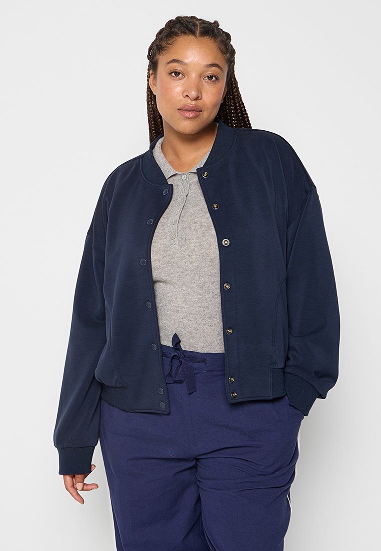 Femme avec les cheveux tressés portant une veste boutonnée bleu marine, un polo gris et un pantalon bleu marine à cordon, se tenant devant un fond clair et uni.