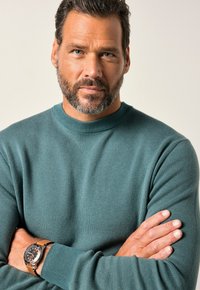 Hombre con barba y ojos azules que lleva un suéter azul verdoso y un reloj en la muñeca, con los brazos cruzados, frente a un fondo beige liso.