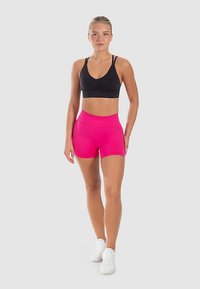 Zwarte sportbeha met een ronde halslijn en roze sportshorts met hoge taille. Model draagt witte sneakers en staat tegen een neutrale achtergrond.