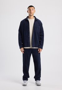 Navy corduroy jacket with button closure and pocket, paired with matching trouser. Light beige t-shirt underneath. White sneakers with gray accents.