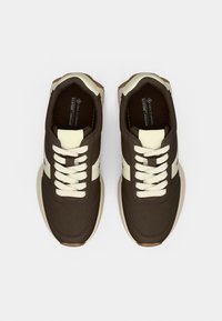 Καφέ vegan αθλητικά παπούτσια με εκρού λεπτομέρειες, με λεία υφή, στρογγυλή μύτη και επίπεδα κορδόνια. Η σόλα είναι μπεζ με ελαφρύ ανάγλυφο πάτημα.