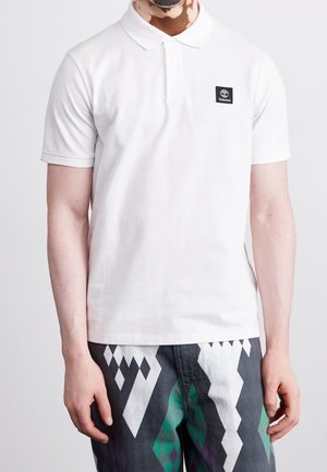 Homme portant un polo Timberland blanc et un pantalon à motifs noir, blanc et vert, debout les bras détendus le long du corps.