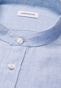 Chemise en lin bleu clair avec col montant et boutons blancs, avec une étiquette de la marque Seidensticker à l’intérieur du col.