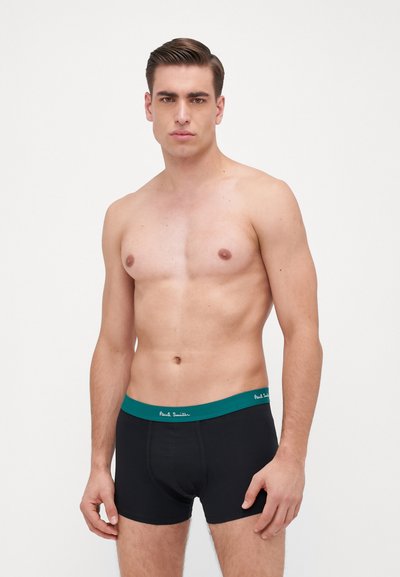 Boxer shorts en coton noir avec une ceinture teal portant du texte vert. Le modèle est torse nu, affichant une expression neutre.