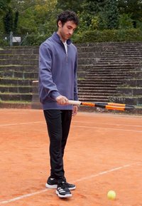 Muž v námorníckej zip-oversize mikine a čiernych nohaviciach drží tenisovú raketu s oranžovými akcentmi na antukovom kurte, pripravený zasiahnuť tenisový loptičku.